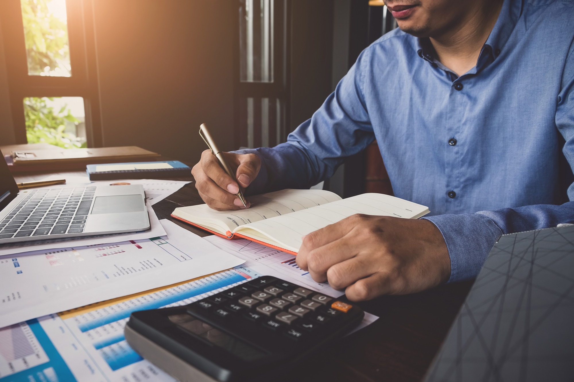 A man writes in a notebook at a desk filled with a laptop, documents, and a calculator, illustrating productivity and planning.