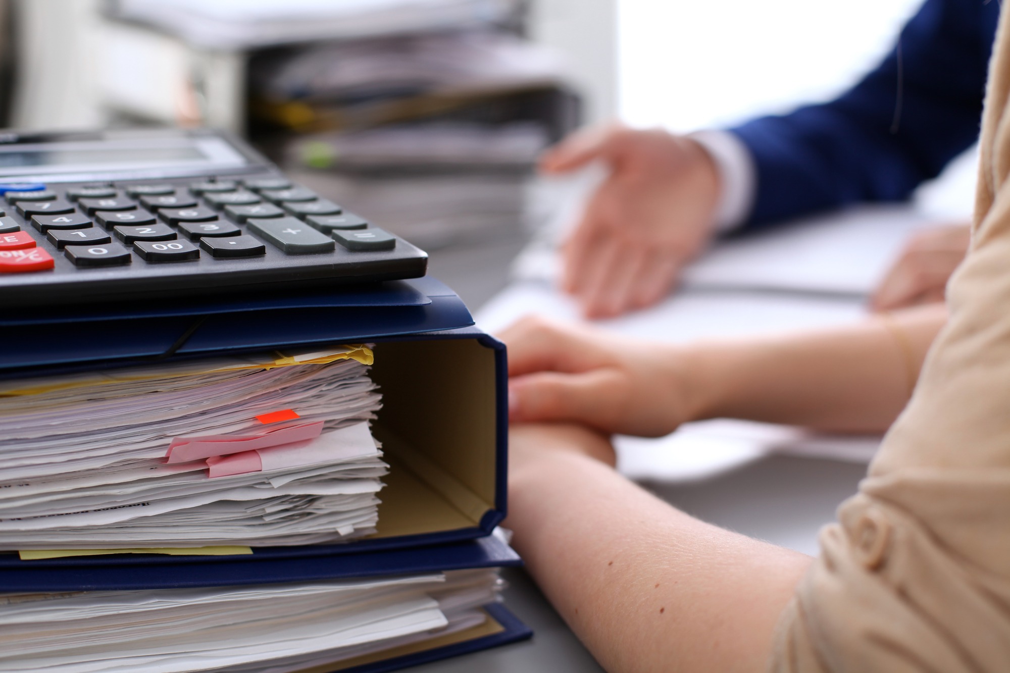 Calculator and stacked papers dominate the workspace, with hands gesturing over documents, suggesting a collaborative discussion or financial review.