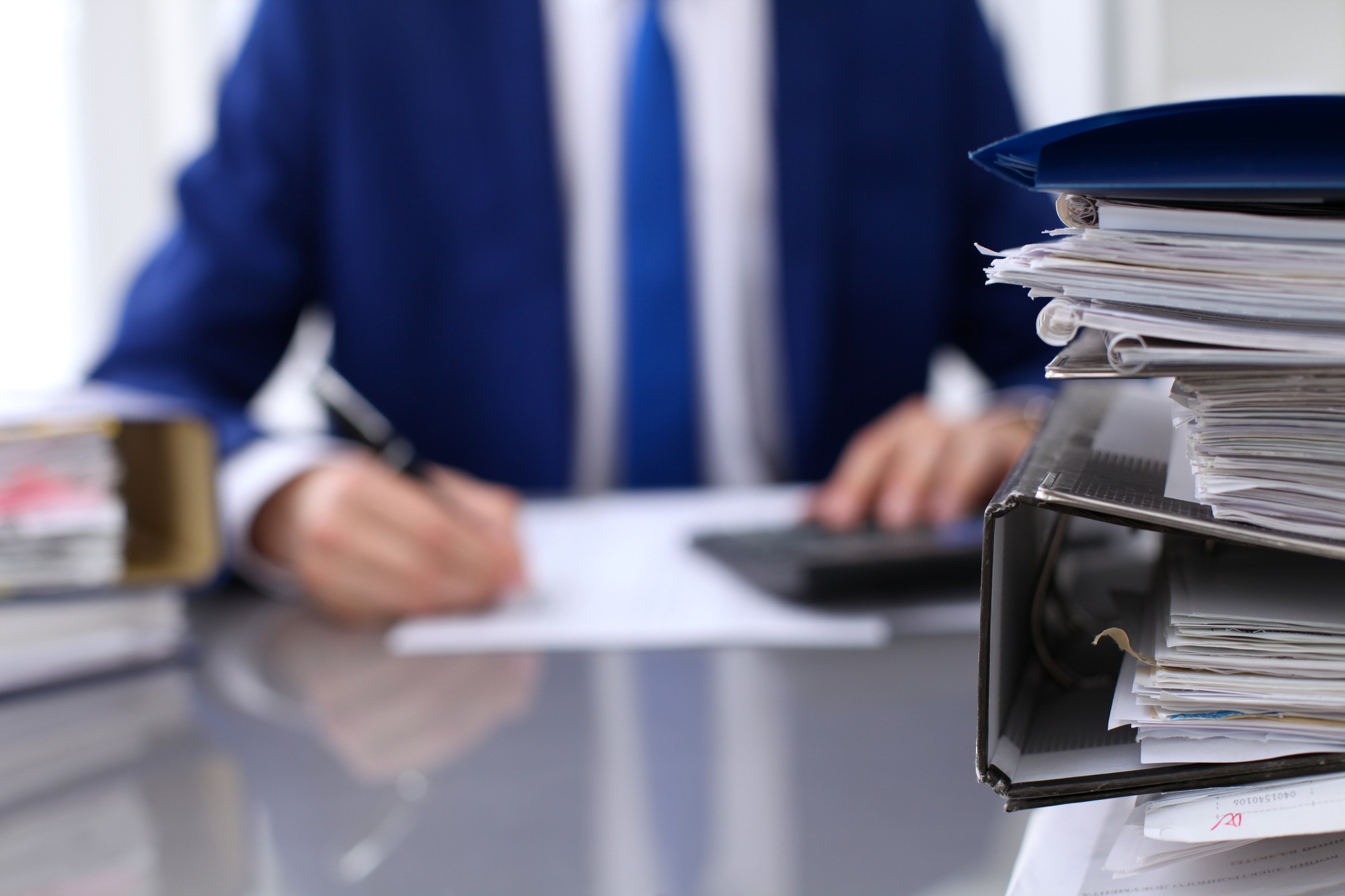 A businessman writes notes while surrounded by stacks of paperwork and a binder